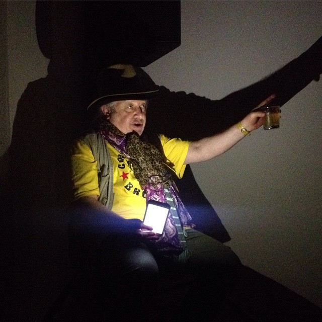 Composer and performer Charlemagne Palestine, holding a mobile phone and drink, a deep shadow behind him.